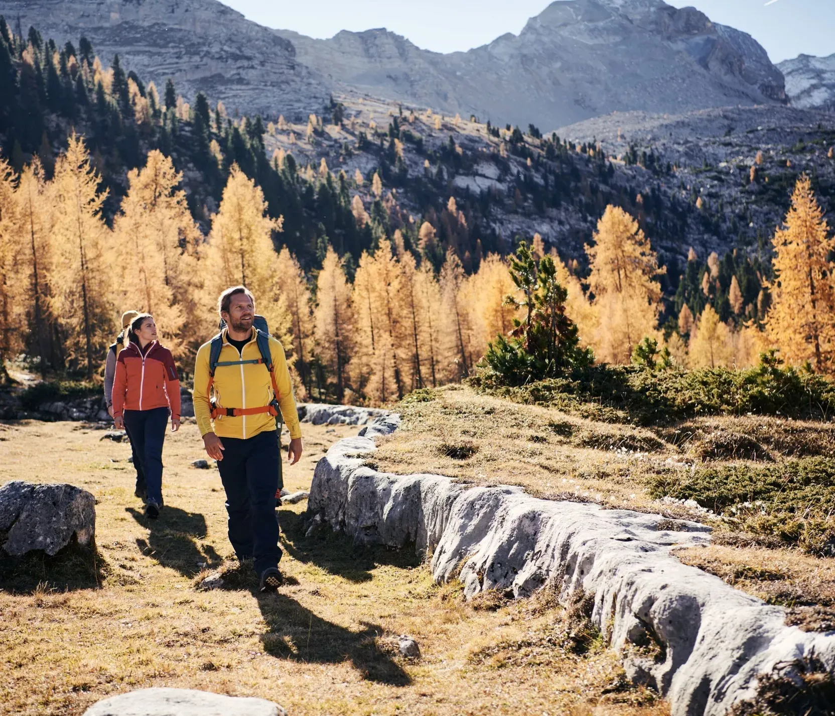 Due escursionisti camminano nella natura tra alberi dorati e montagne, con un cielo chiaro e paesaggi di montagne sovrastanti.