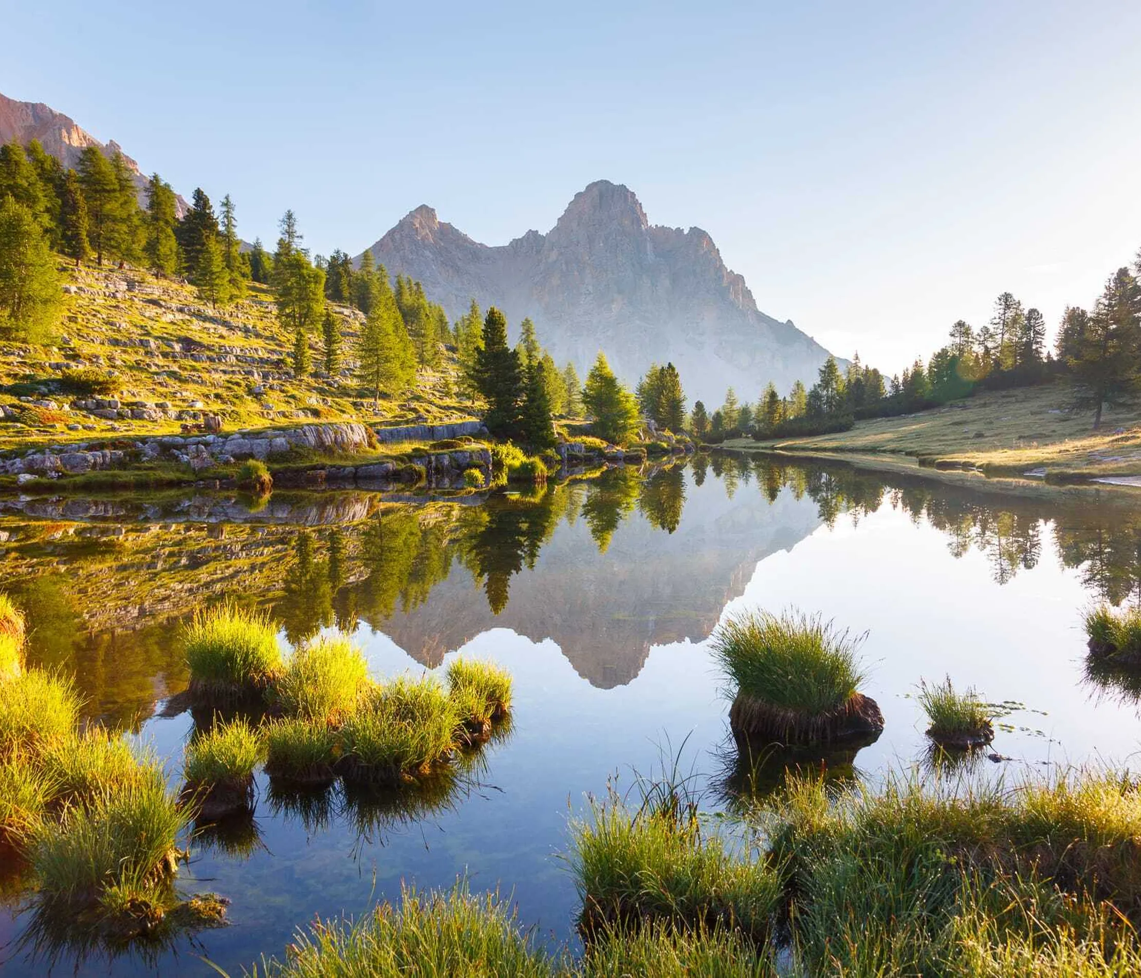 Lago riflettente circondato da alberi verdi e montagne rocciose sullo Sfondo, con atmosfera serena e illuminata dalla luce del mattino o del tramonto.