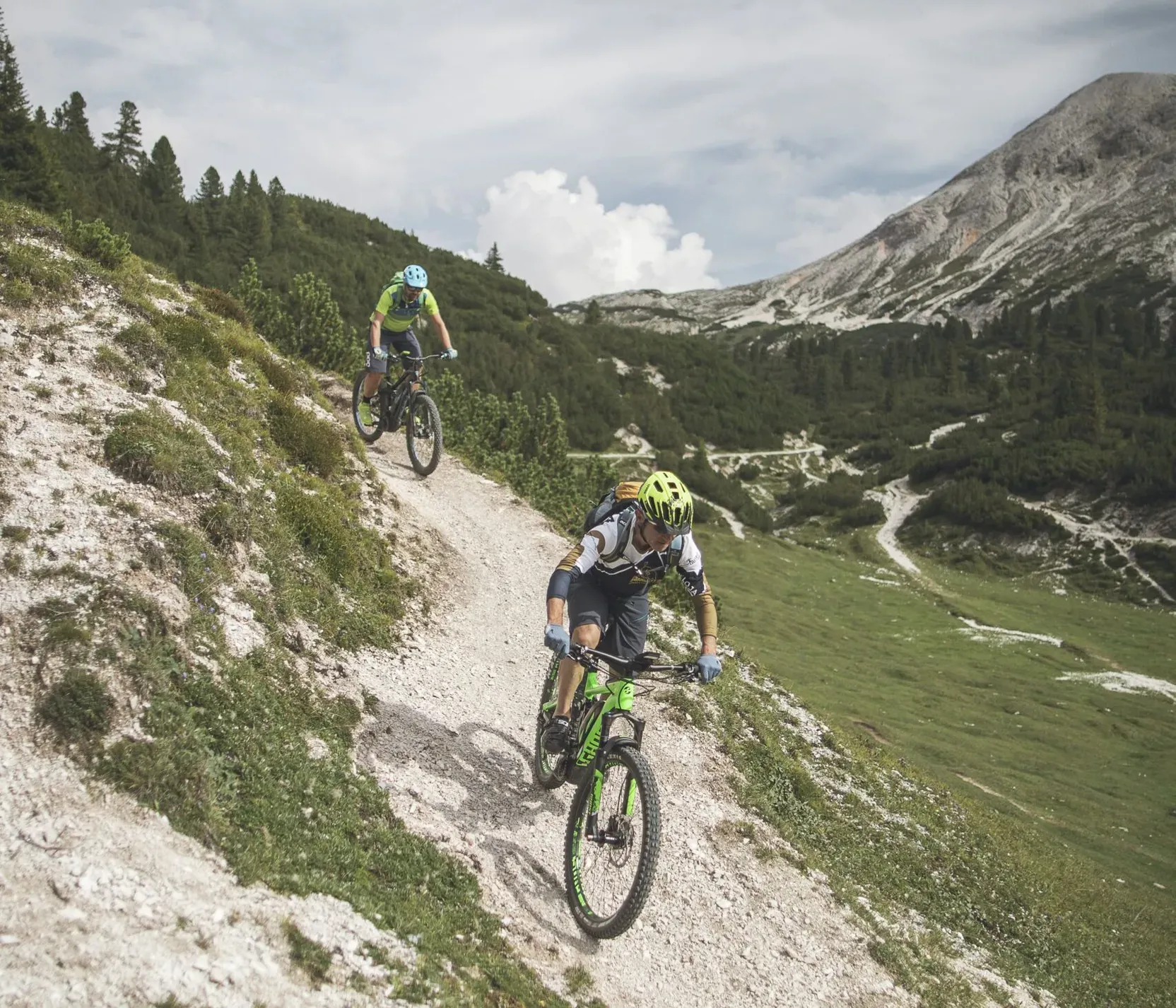 Due mountain bikers descend a rocky trail in the Dolomites, with green vegetation, a mountain in the background, and a partly cloudy sky above.