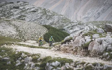Zwei Mountainbiker fahren auf einem schmalen Weg durch eine alpine Landschaft mit grünen Hügeln und felsigen Bergen im Hintergrund.
