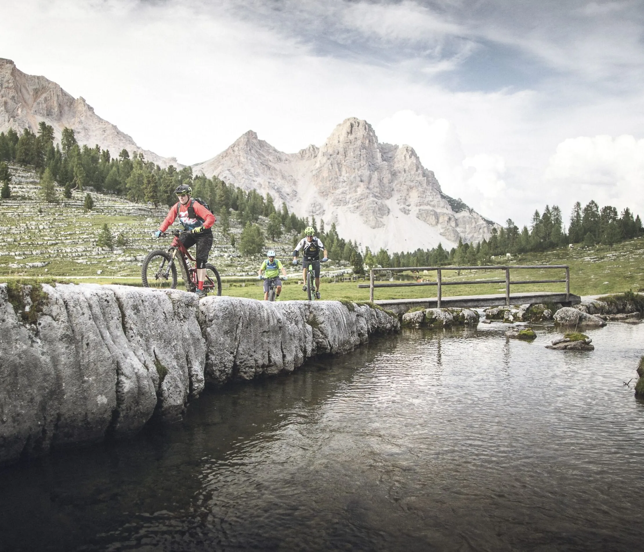 Escursionisti in mountain bike attraversano un fiume tra le Alpi Dolomiti di Alta Badia, con montagne e cielo nuvoloso sullo sfondo.