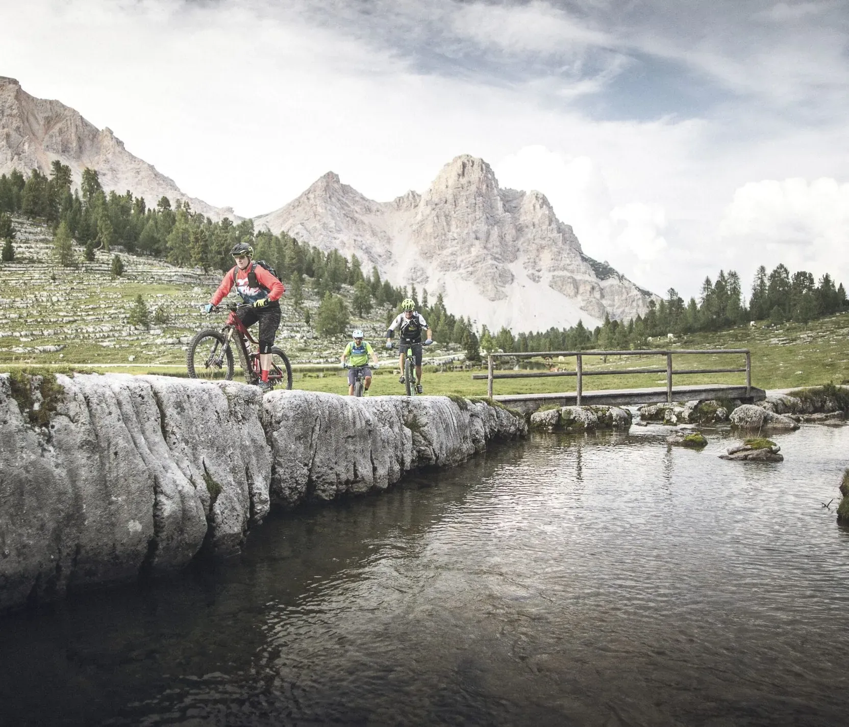 Drei Mountainbiker fahren über einen Felsen neben einem Fluss, umgeben von Bergen und grünen Wiesen unter einem bewölkten Himmel.