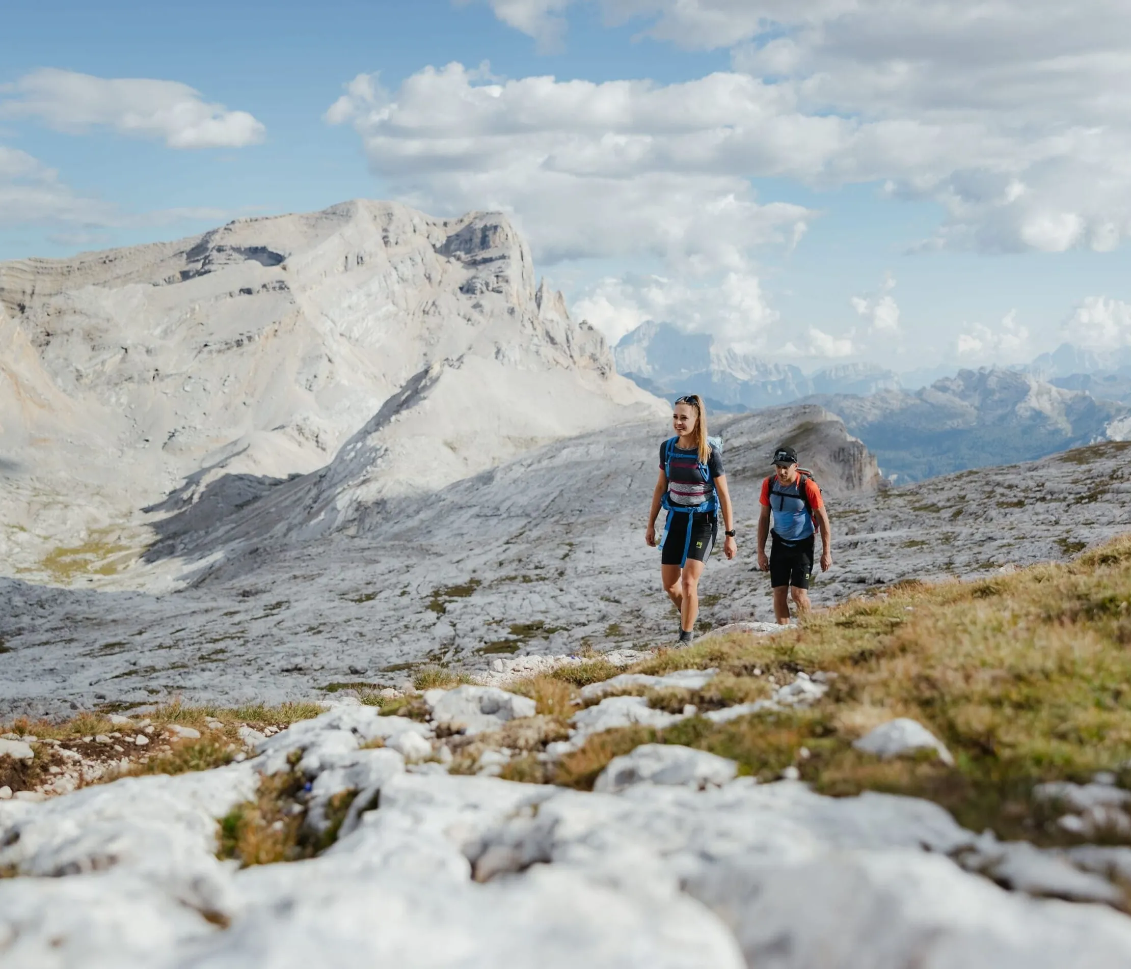Zwei Wanderer auf einem felsigen Pfad in den Dolomiten, umgeben von imposanten, grauen Bergen unter einem Himmel mit weißen Wolken.