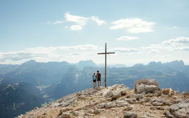 Zwei Wanderer auf einem Berggipfel neben einem Holzkreuz mit Blick auf die Dolomiten, Wolken im blauen Himmel über der atemberaubenden Landschaft.
