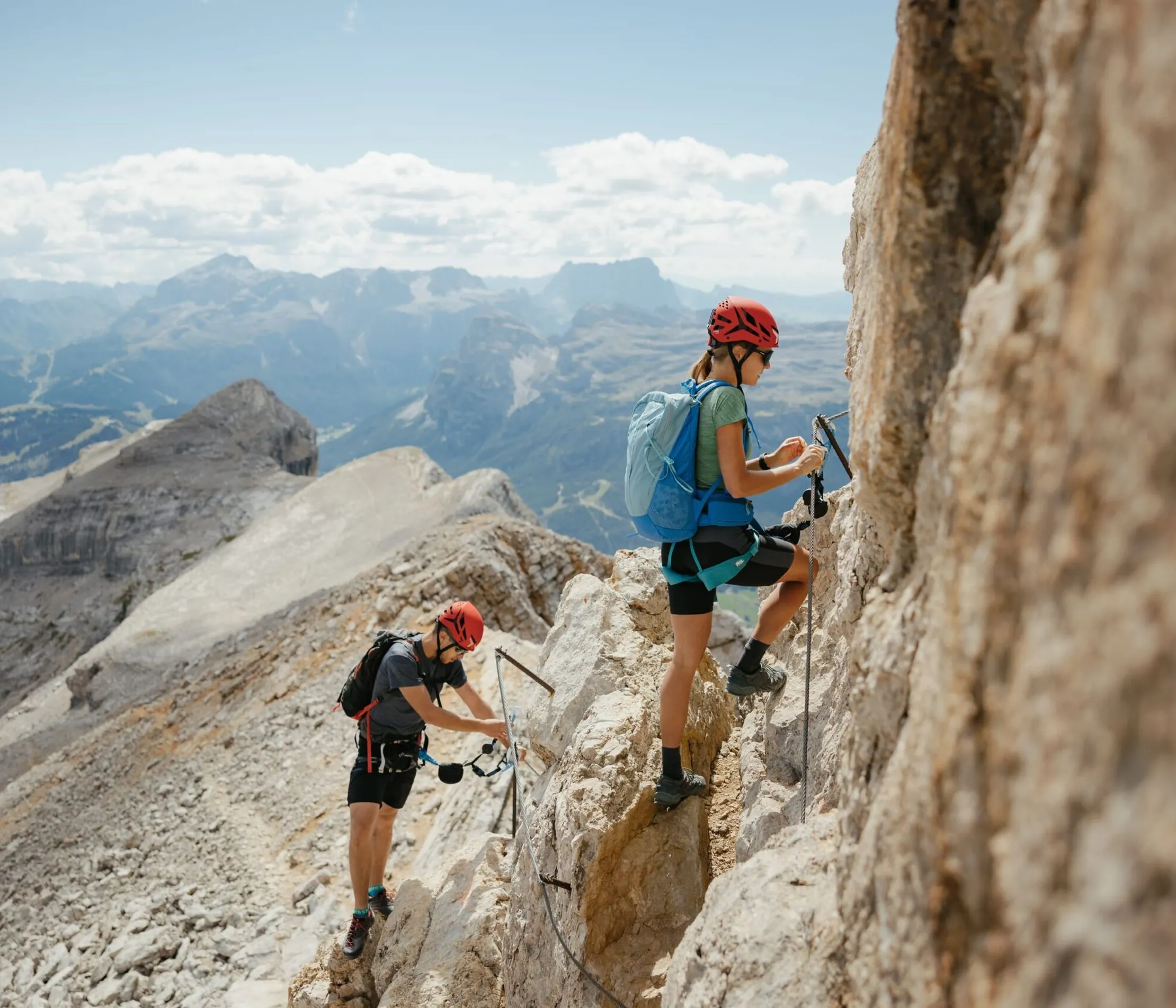 Two climbers with helmets and backpacks ascending a rocky mountain trail in the Dolomites, with scenic peaks and clouds in the background.