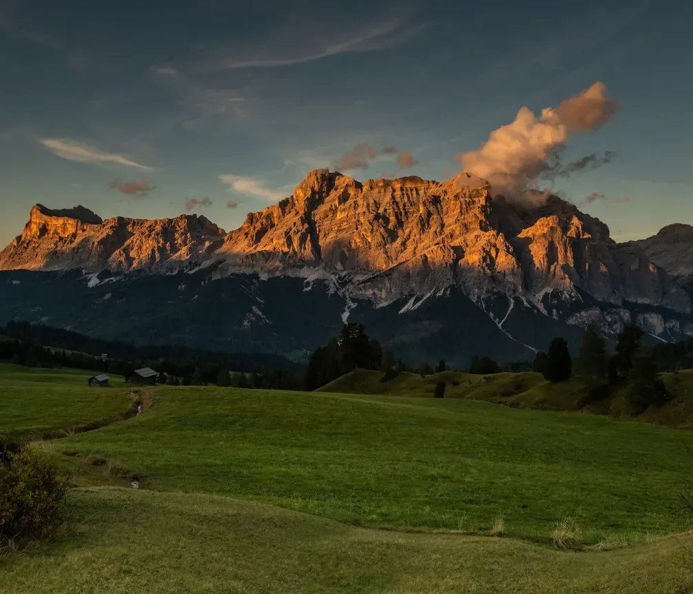 Muntagne Dolomiti al tramonto, con cime rocciose illuminate dal sole e un cielo con alcune nuvole, fronte un prato verde con alberi e piccoli edifici.