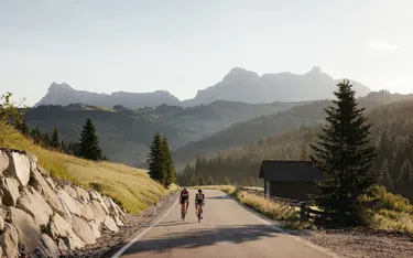Zwei Radfahrer fahren auf einer bergigen Straße durch eine grüne Landschaft, umgeben von Hügeln, Tannenbäumen und imposanten Bergen im Hintergrund.