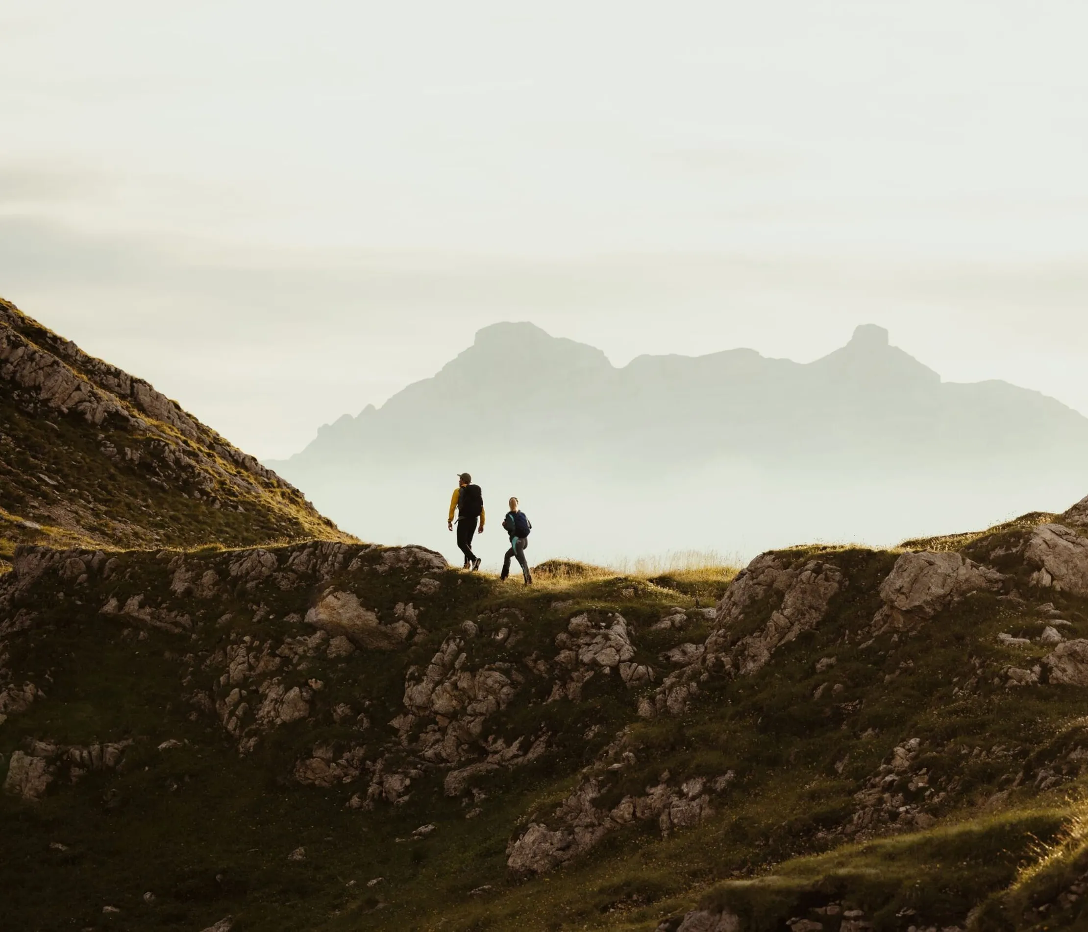 Due escursionisti camminano su un sentiero tra le montagne delle Dolomiti, con un paesaggio montano sfocato sullo sfondo in una luce soffusa.