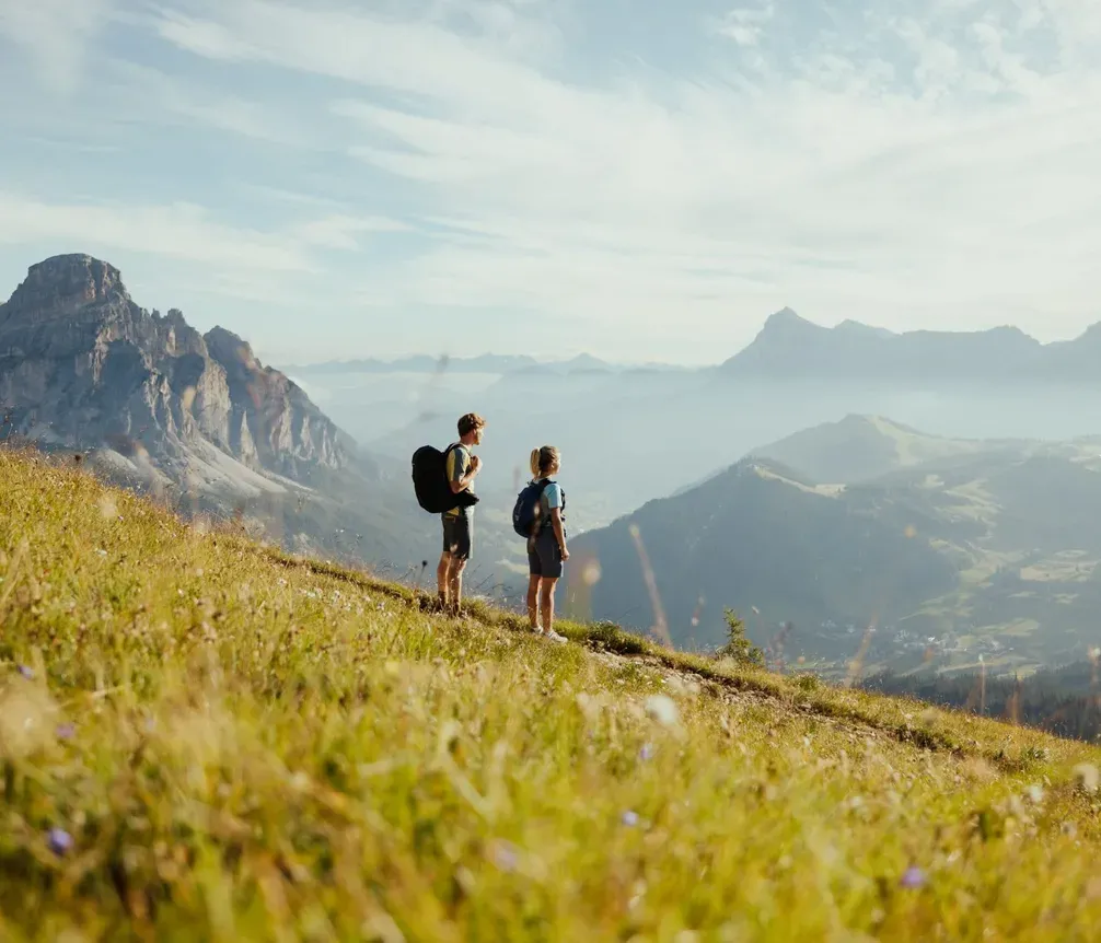 Due giovani escursionisti camminano su un prato in Alta Badia con montagne sullo sfondo, cielo nuvoloso e atmosfera serena.