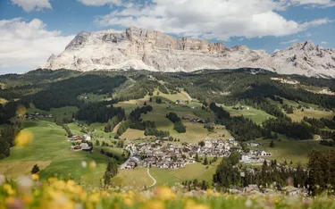 Dorf in den Dolomiten mit grünem Hügel, Wäldern und Berggipfeln im Hintergrund, unter einem bewölkten Himmel.