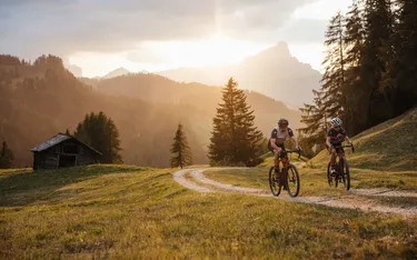 Zwei Mountainbiker fahren auf einem schmalen Weg durch eine grüne Hügellandschaft in den Dolomiten, umgeben von Bäumen, mit Bergen im Hintergrund bei sunset.