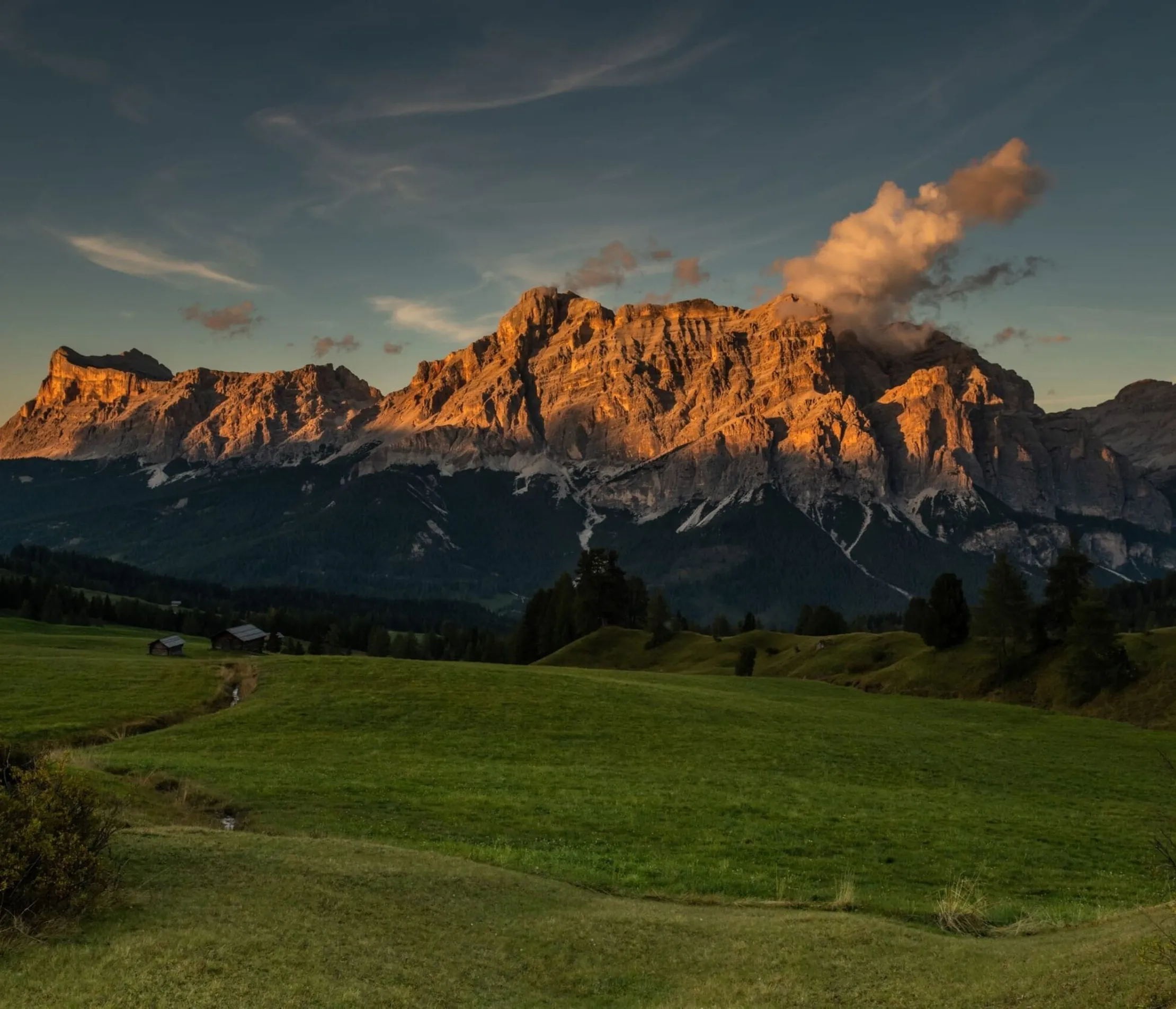 Bergmassiv bei Sonnenuntergang mit grünen Wiesen im Vordergrund und kleinen Hütten, Wolken ziehen um die Gipfel.