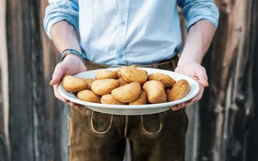 Person hält einen weißen Teller mit goldgelben, gebackenen Krapfen vor einem Holzzaun, getragen in einer blau-weißen Hemd- und braunen Lederhosen-Kleidung.