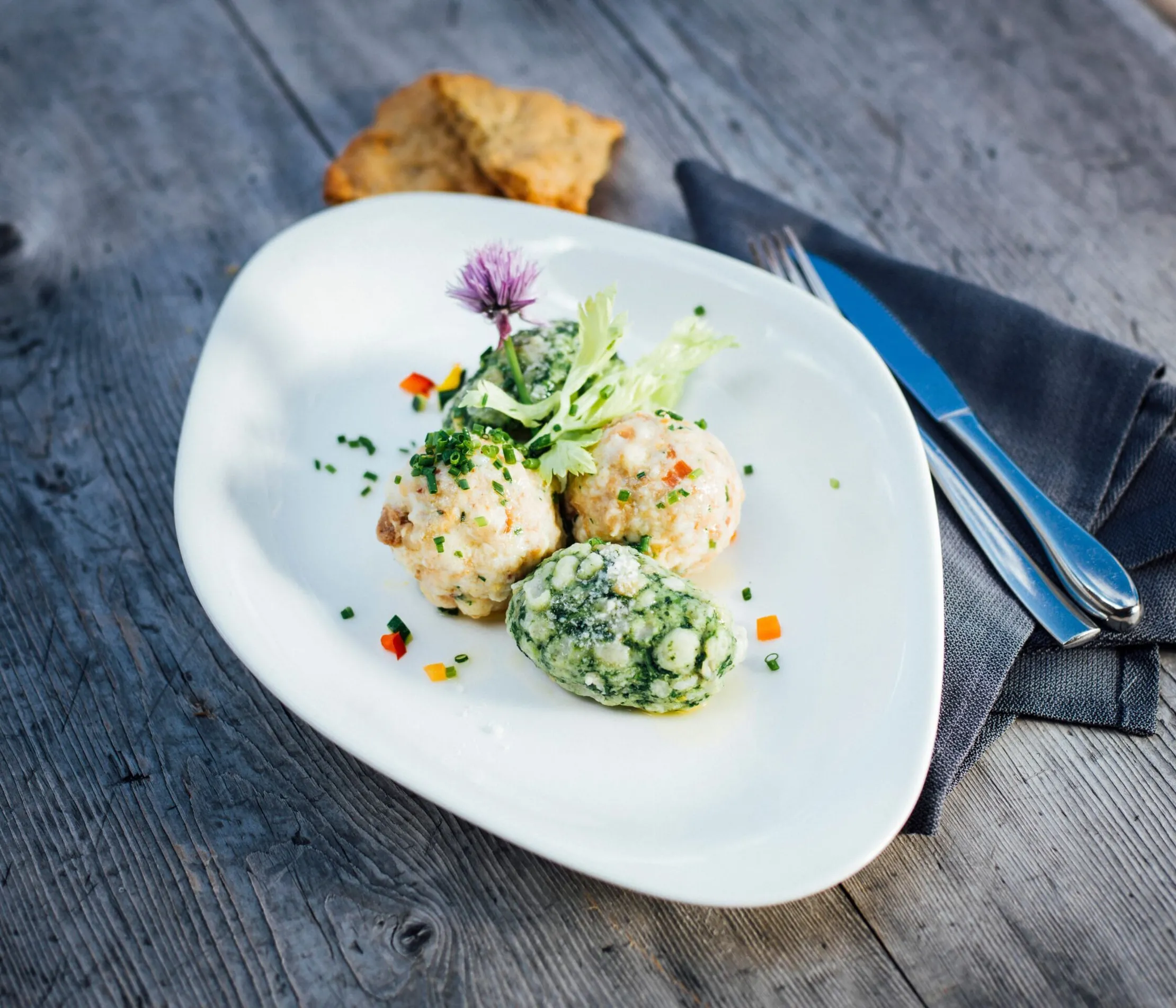 Polpette di pesce con verde e fiori freschi su un piatto bianco, posato su un tavolo di legno grezzo, con posate e tovagliolo nero accanto.