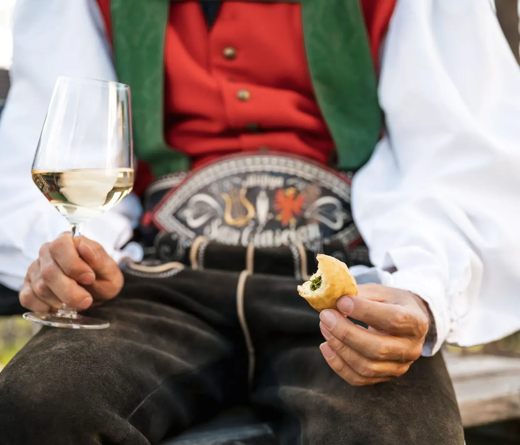 Una persona in abiti tradizionali seduta su una panca, tiene un bicchiere di vino bianco e un pezzo di cibo in mano, con uno sfondo naturale e luminoso.