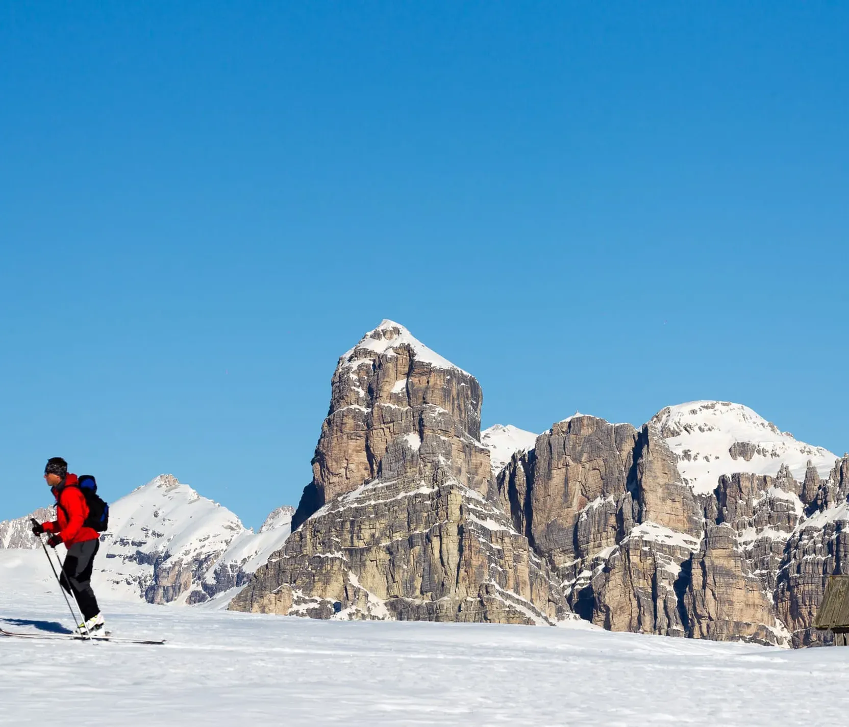 Skifahrer in rot und schwarz mit Rucksack auf verschneiter Landschaft vor den Dolomiten im blauen Himmel.
