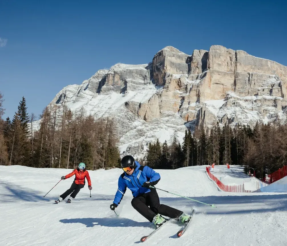 Due sciatori in pista sulla neve, con montagne e boschi sullo sfondo, sotto un cielo azzurro in una località sciistica delle Dolomiti.