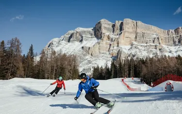 Zwei Skifahrer in winterlicher Landschaft mit Snowboard und Bergen im Hintergrund, klare blauer Himmel, schneebedeckte Pisten und Tannen.
