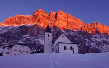 Eine weiße Kirche mit dunklem Dach und Glockenturm vor einer rot-orange leuchtenden Felswand im Dolomiten-Gebirge bei Sonnenuntergang, umgeben von verschneitem Gelände.