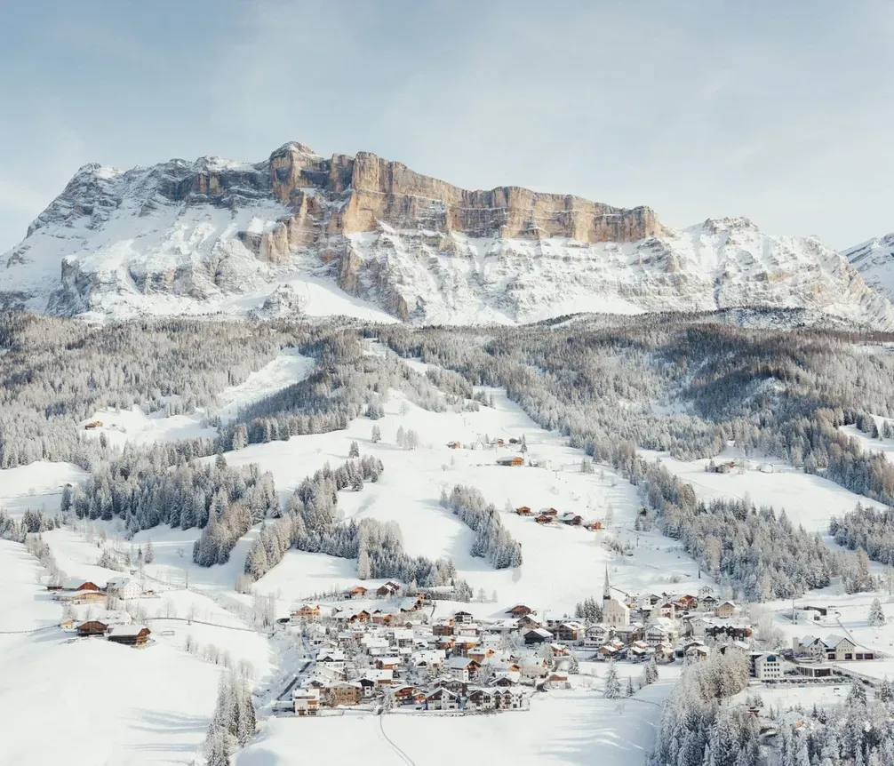 Paesaggio invernale con un piccolo villaggio innevato ai piedi di montagne rocciose e alberi ricoperti di neve, sotto un cielo chiaro.