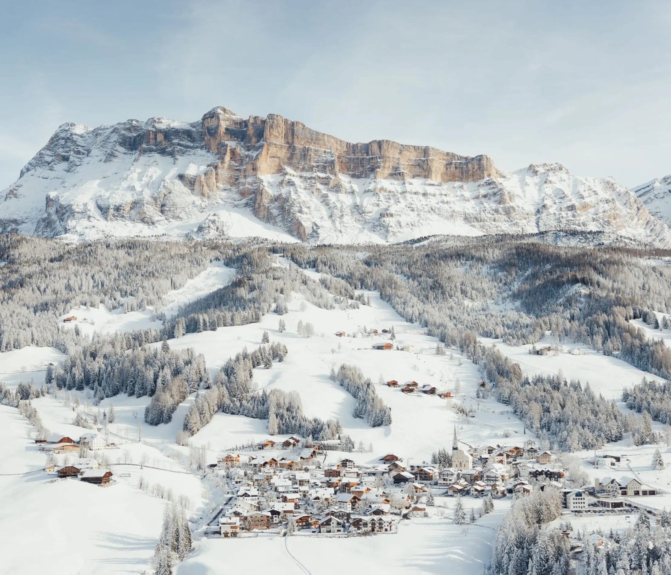 Schneebedeckte Berge in den Dolomiten mit einem kleinen Dorf am Fuße, umgeben von verschneiten Wäldern und Hängen.