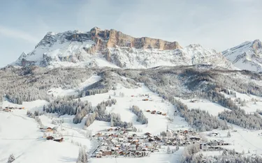 Schneebedeckte Berge in den Dolomiten mit einem kleinen Dorf am Fuße, umgeben von verschneiten Wäldern und Hängen.