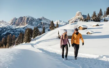 Zwei Menschen wandern in Schneelandschaft in Alta Badia, Dolomiten, mit Berghütten und Gipfeln im Hintergrund, bei klarem Himmel.