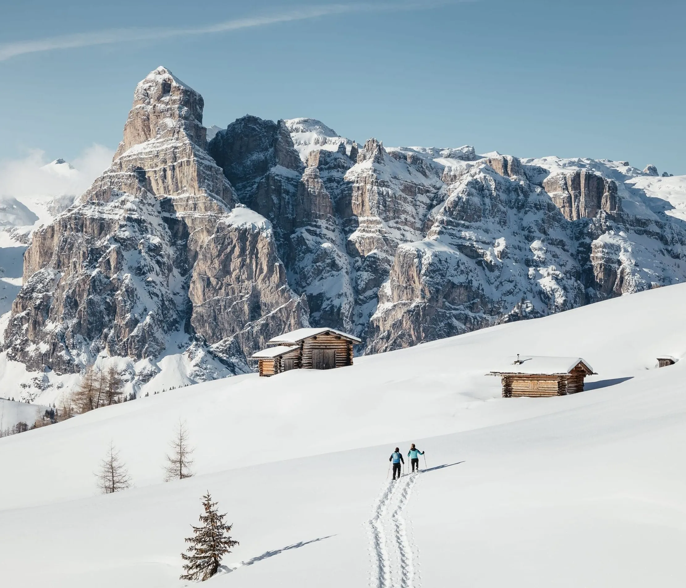 Zwei Personen wandern im Schnee vor Berghütten mit den Dolomiten im Hintergrund, unter klarem Himmel in Alta Badia, Dolomiten.