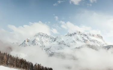 Schneebedeckte Berge in den Dolomiten, umgeben von Wolken und Nebel, mit einem klaren blauen Himmel im Hintergrund.