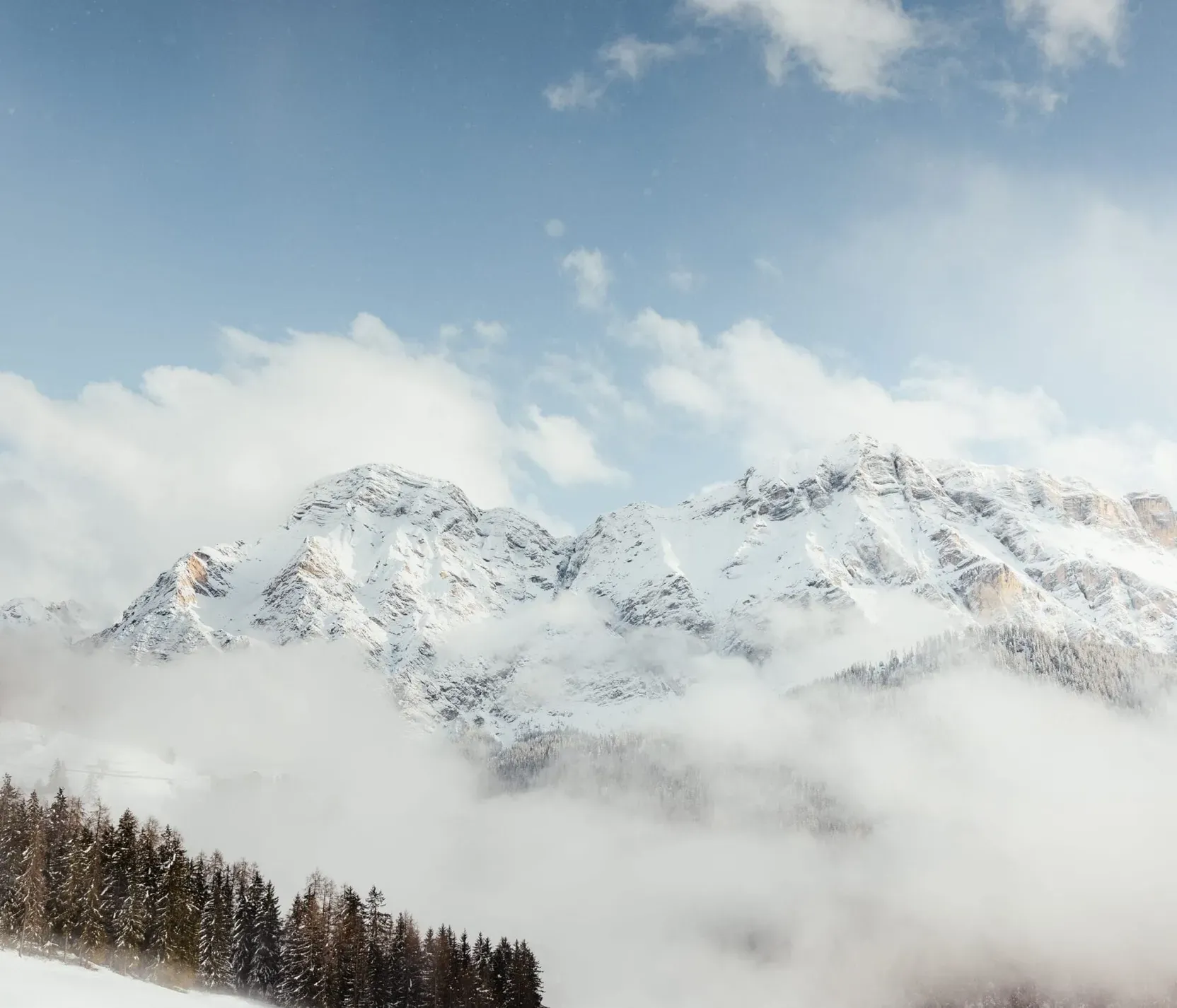 Schneebedeckte Berge in den Dolomiten, umgeben von Wolken und Nebel, mit einem klaren blauen Himmel im Hintergrund.