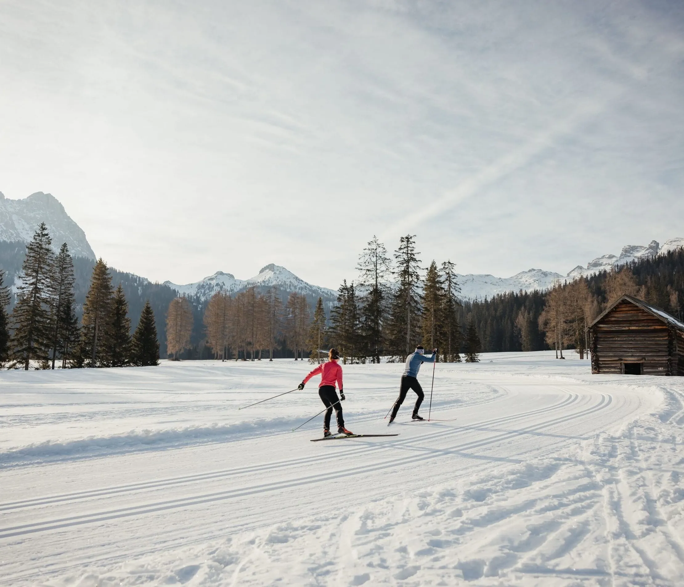 Zwei Skifahrer auf Schneefeld vor Baumgruppe und Bergen in Alta Badia, Dolomiten, sonniger Tag, im Hintergrund eine kleine Holzhütte.