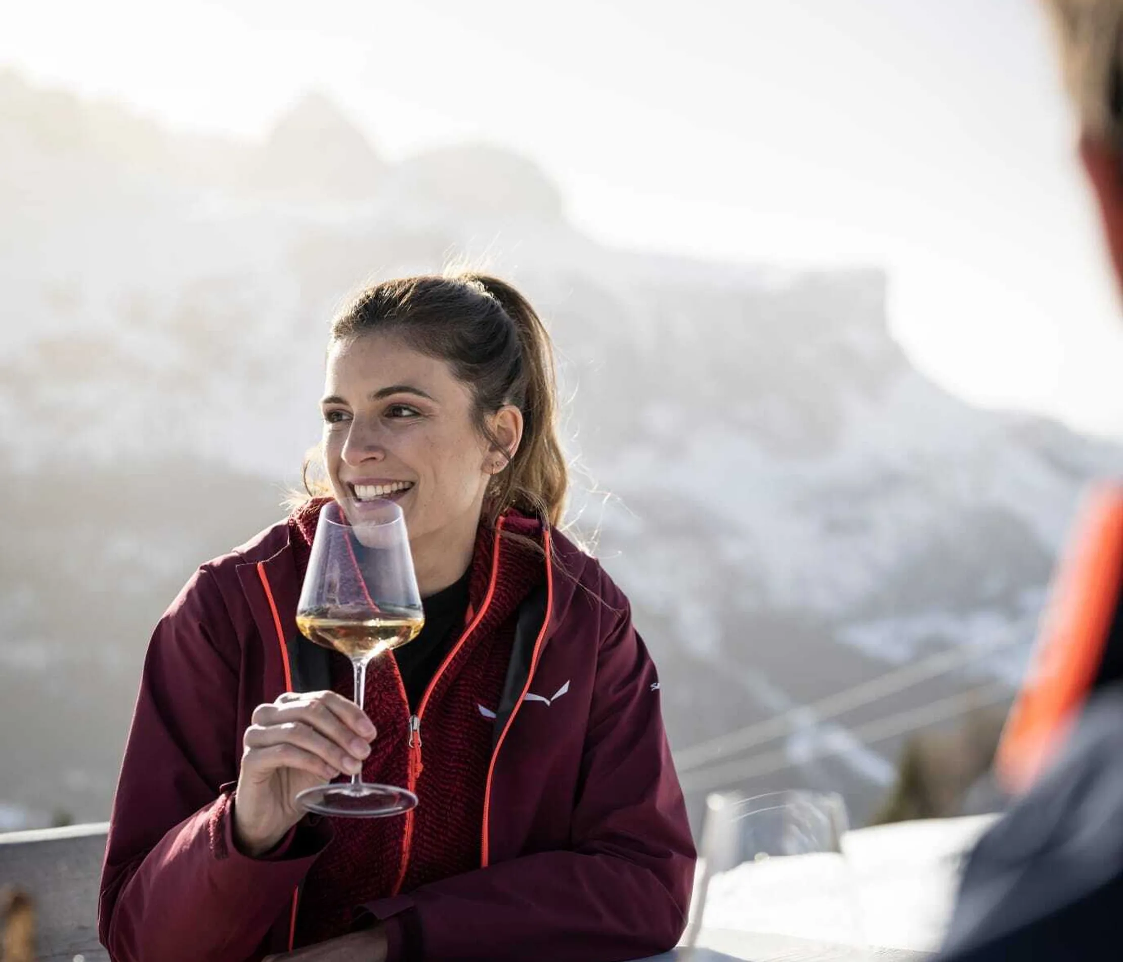 Frau mit braunem Haar in Zopf, trägt rote Jacke, sitzt draußen mit Wein, lächelt, im Bergpanorama der Dolomiten, beim Essen am Balkon.