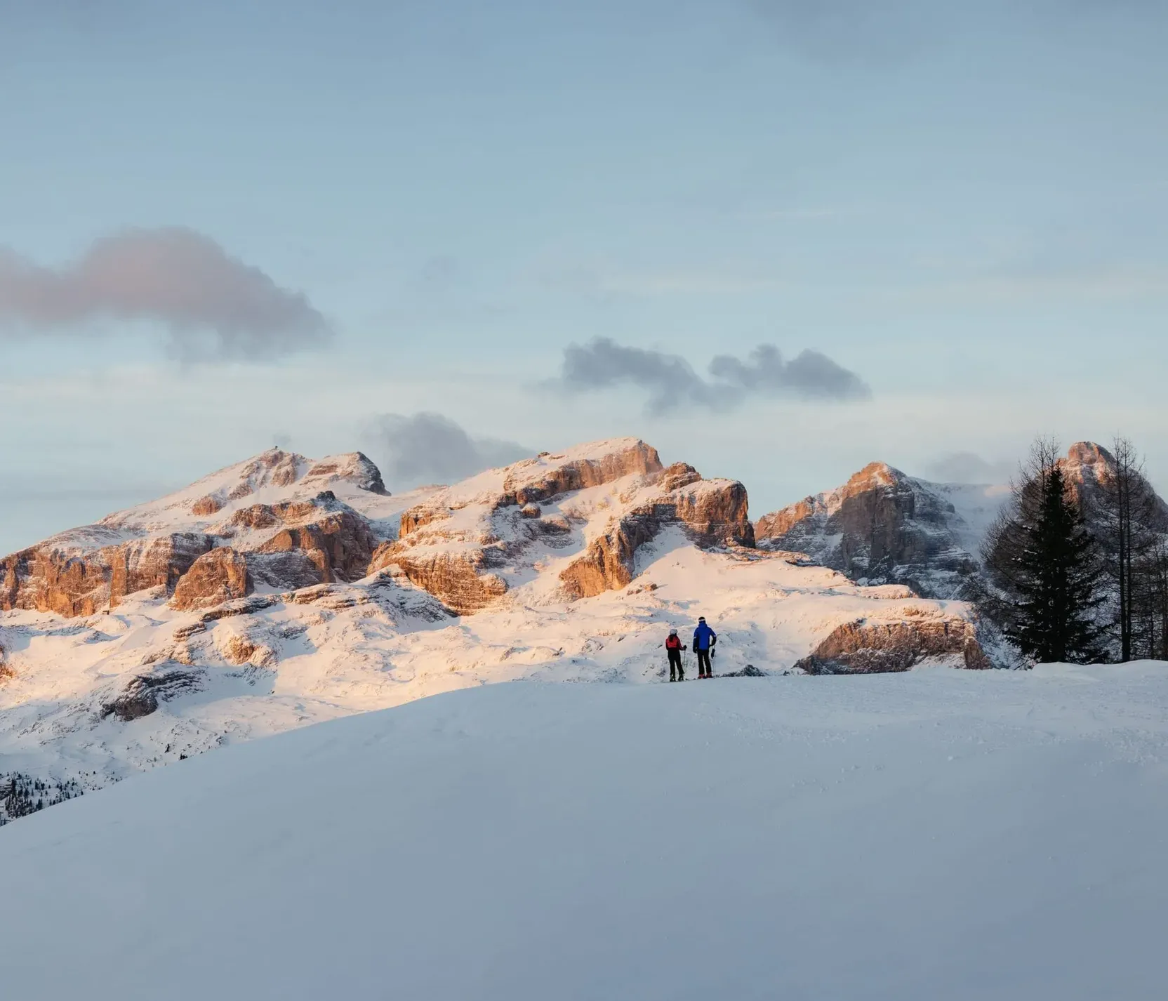 Two hikers stand on a snowy hill with the rugged, snow-covered Dolomite mountains in the background under a partly cloudy sky.