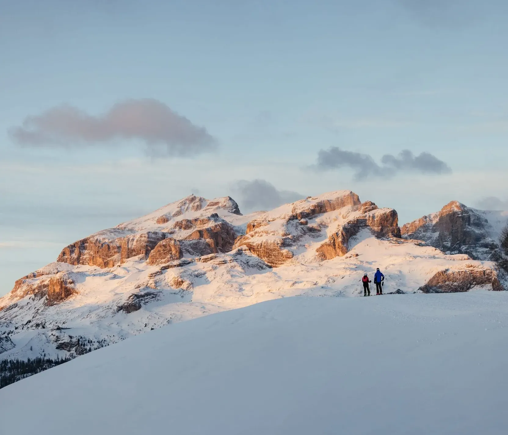 Zwei Personen wandern in verschneiter Berglandschaft mit Felsen und Nadelbäumen, bei Sonnenaufgang in der Dolomitenregion Alta Badia.