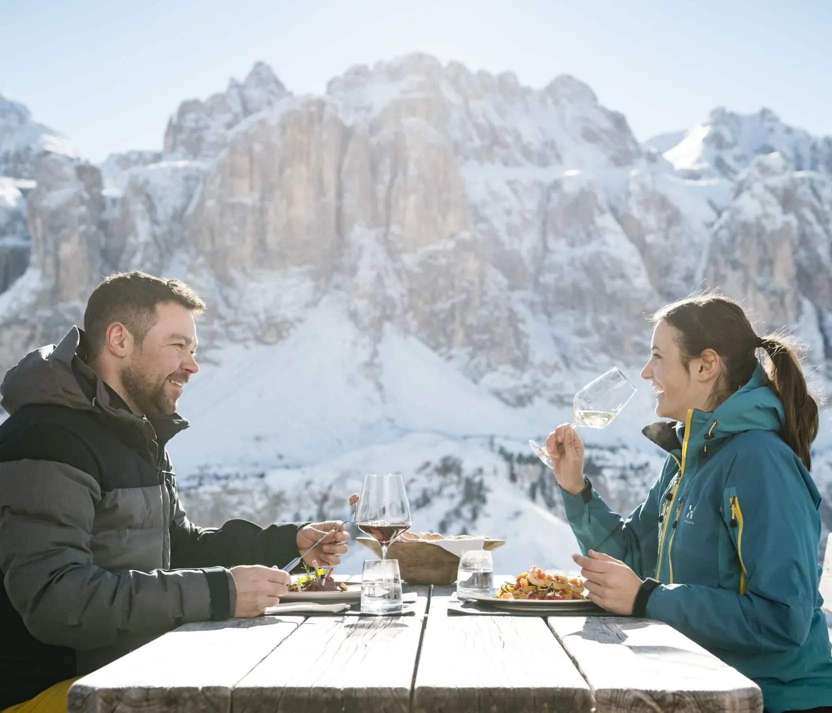 Zwei Personen beim Essen im Freien mit Blick auf verschneite Dolomitenberge, tragen Wintersportkleidung und genießen Wein und Pizza.