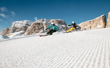 Zwei Skifahrer in Skiunterricht in Alta Badia, Dolomiten, auf perfekt präparierter Piste vor beeindruckender Bergkulisse bei schönem Wetter.