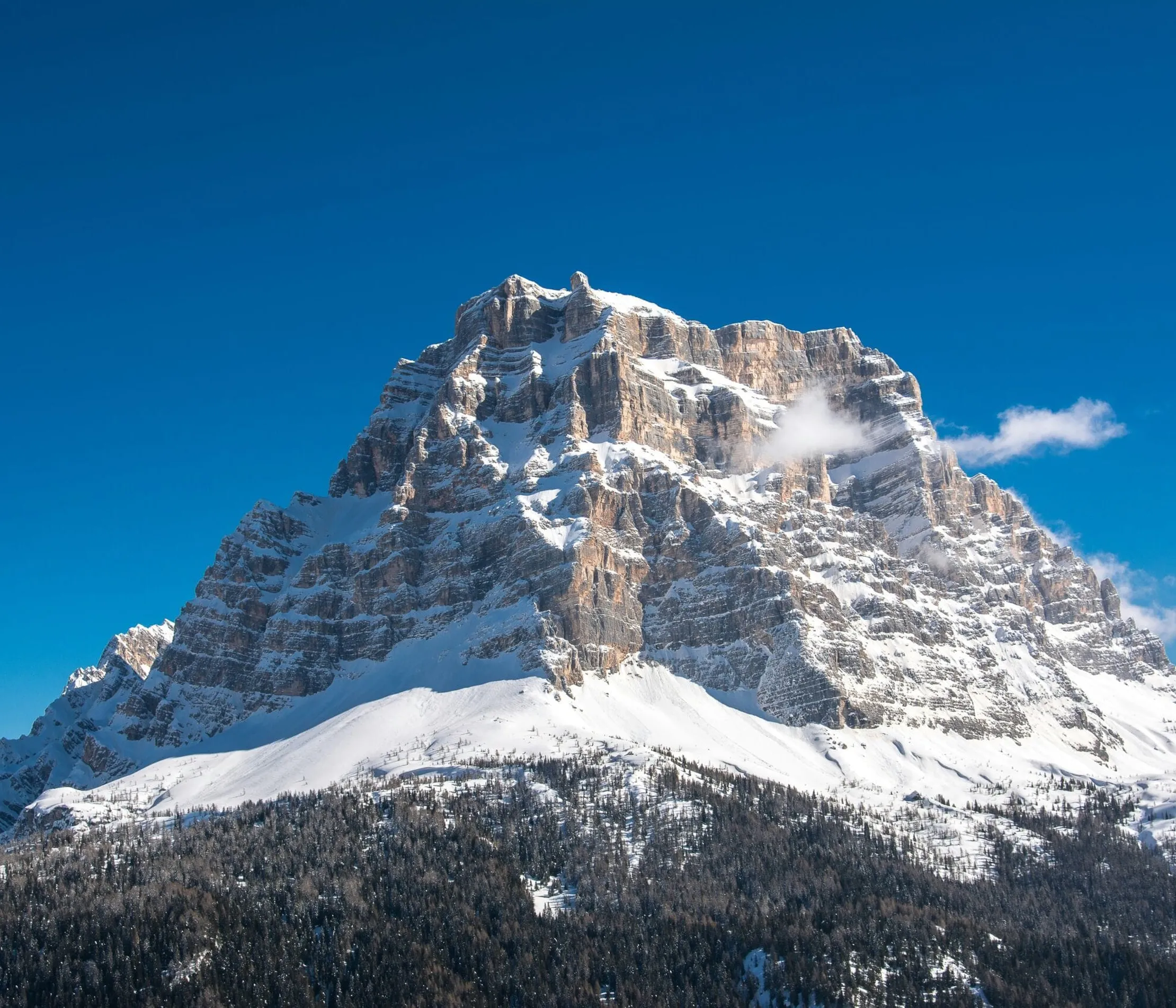 Maestosa montagna imbiancata di neve con cielo azzurro, circondata da foresta e vette rocciose in alta quota nelle Dolomiti.