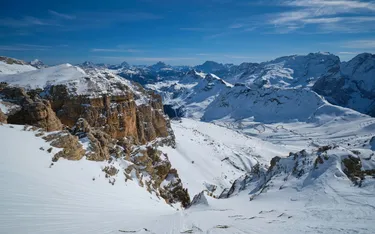 Breiter Blick über verschneite Bergen und Täler in den Dolomiten, mit einem klaren blauen Himmel und einigen Wolken im Hintergrund.