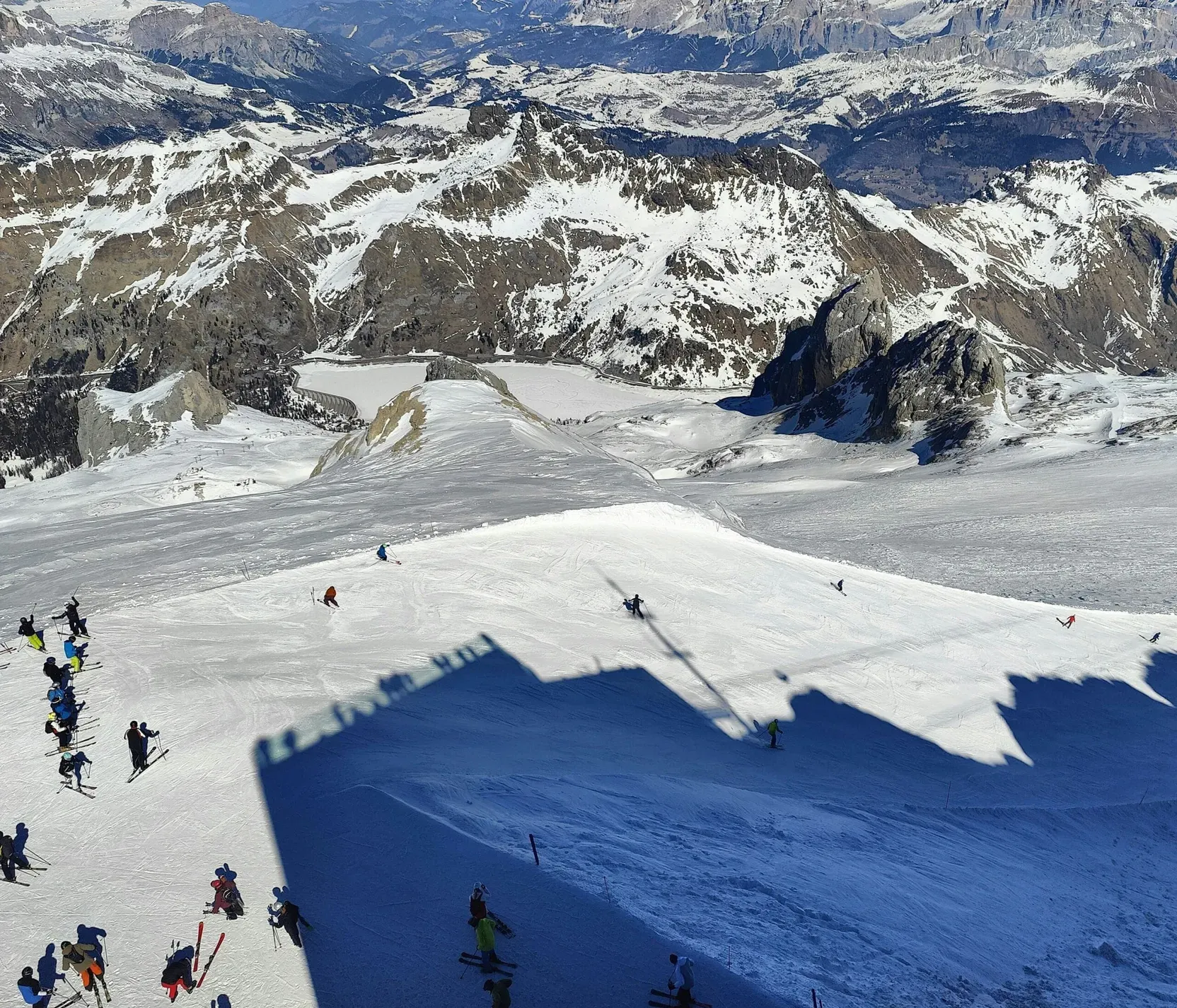 Sciatori e snowboarder sulla neve, con montagne innevate e cime rocciose sullo sfondo nelle Dolomiti. In secondo piano, l'ombra di una struttura sul pendio.