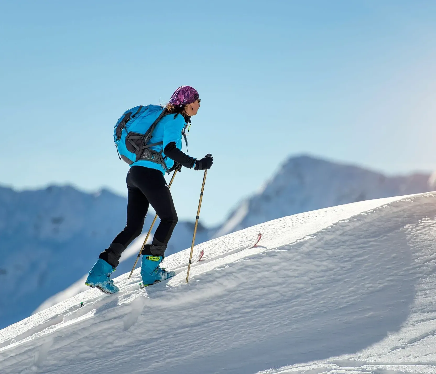 Frau beim Skitourenaufstieg in den schneebedeckten Dolomiten bei Sonne, mit blauem Rucksack und pinkem Stirnband.