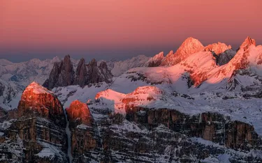 Snow-covered mountain peaks at sunset in Alta Badia, Dolomites, with pink and orange hues illuminating the rugged terrain.